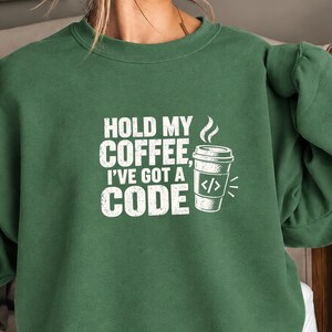 May include: A forest green crewneck sweatshirt featuring white screen-printed text that reads "HOLD MY COFFEE, I'VE GOT A CODE" alongside a graphic of a disposable coffee cup with a programming code bracket symbol on the front of the cup.