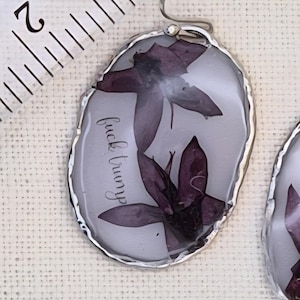May include: An oval-shaped pendant featuring two pressed purple flowers encased in clear resin with a silver-toned metal border. The text "fuck trump" is printed in black script on the surface. A ruler shows the pendant is approximately 1.5 inches tall.