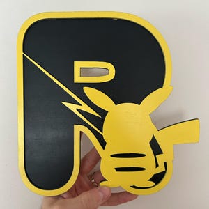 May include: A black and yellow wooden letter R with a Pikachu silhouette inside. The letter is outlined in yellow and the Pikachu is facing the right side of the letter.