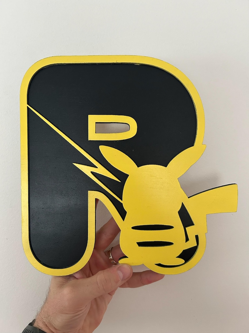May include: A black and yellow wooden letter R with a Pikachu silhouette inside. The letter is outlined in yellow and the Pikachu is facing the right side of the letter.