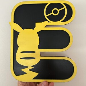 May include: A wooden letter E with a yellow outline and a black interior. The letter E is decorated with a yellow Pikachu character and a black Pokeball.