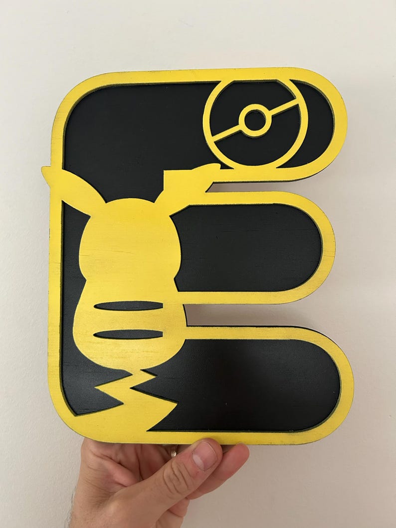 May include: A wooden letter E with a yellow outline and a black interior. The letter E is decorated with a yellow Pikachu character and a black Pokeball.