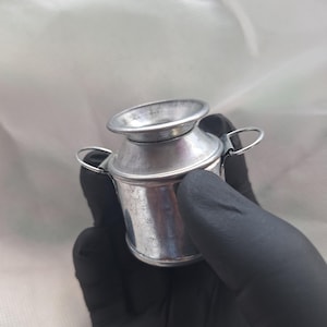 May include: A small, silver-colored metal milk can with a flared top and two small handles. The miniature can is held in a gloved hand against a white background. The can has a vintage, rustic appearance.