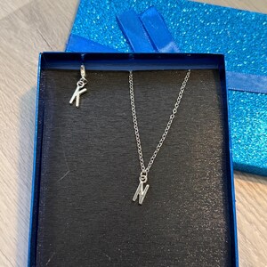 Puede incluir: Un collar plateado con un colgante con la letra "N" y un charm "K" separado, presentado en una caja de regalo azul con tapa a juego. La caja tiene un acabado brillante y una cinta azul.