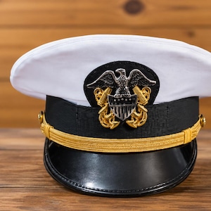 Könnte beinhalten: Eine weiß gekrönte Dienstmütze mit schwarzem Band, schwarzem Schirm und goldenem Kinnriemen. In der Mitte befindet sich ein metallisches Emblem, das einen silbernen Adler über einem Schild zeigt, flankiert von zwei goldenen gekreuzten Ankern.