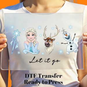May include: A clear, rectangular DTF transfer featuring Elsa, Olaf, Sven, and a snowflake wand. The phrase "Let it go" is written in cursive. The text "DTF Transfer Ready to Press" is at the bottom.