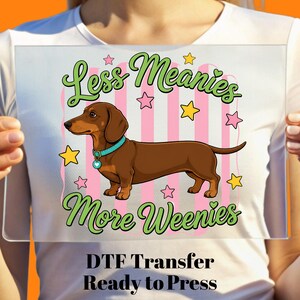 Peut inclure: Un transfert DTF avec un teckel de dessin animé avec un collier bleu, entouré d'étoiles et de rayures roses. Le texte dit "Less Meanies More Weenies" en vert. En dessous, il est écrit "DTF Transfer Ready to Press."