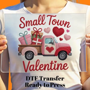 May include: A clear rectangular DTF transfer featuring a pink truck with a heart on the door, carrying gifts and surrounded by hearts. The text reads "Small Town Valentine" and "Ready to Press."