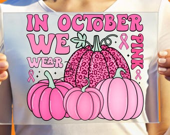 May include: A white t-shirt featuring a graphic design with three pumpkins in shades of pink. The largest pumpkin has a pink leopard print. The text reads IN OCTOBER WE WEAR PINK, accompanied by three small pink awareness ribbons and stars.