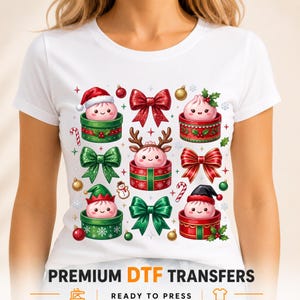 May include: A white t-shirt featuring a graphic print of four cute pink dumplings in gift boxes, decorated with red and green bows, a Santa hat, reindeer antlers, and an elf hat, surrounded by small Christmas ornaments and candy canes.