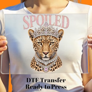 May include: A DTF transfer featuring a leopard head wearing a jewelled crown and a matching diamond necklace. Above the leopard, the word SPOILED is printed in a pink glitter texture. The design is displayed on a clear sheet held in front of a white shirt.