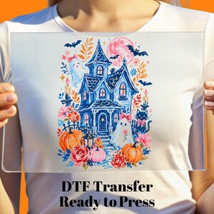 May include: A white t-shirt featuring a colorful graphic print of a blue haunted house, two ghosts with pink bows, a black cat, orange pumpkins, red flowers, bats, and a pink moon. The text DTF Transfer Ready to Press is printed below the graphic.