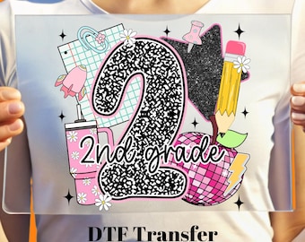 May include: A DTF transfer featuring a large number "2" with a black and white pattern, the words "2nd grade", and school-themed graphics like a pencil, apple, and notebook. The design is in pink, black, and yellow.
