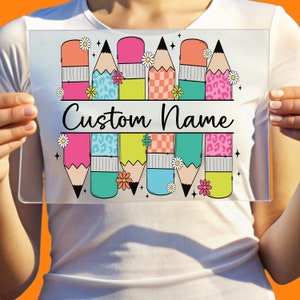 May include: A personalized glass cutting board with a colorful design of pencils, flowers, and stars. The text "Custom Name" is printed in black on the board.