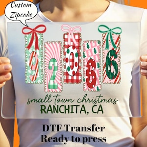 May include: A clear rectangular DTF transfer with the text "small town christmas RANCHITA, CA". The design features the numbers "92066" in a Christmas theme with bows and patterns. The text "DTF Transfer Ready to press" is also included.