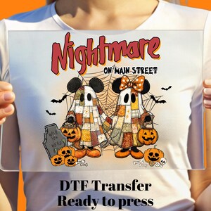 May include: A DTF transfer featuring a Halloween design with the text "Nightmare on Main Street." The image includes two ghost-like figures with mouse ears, holding jack-o'-lanterns. Additional pumpkins, bats, and a tombstone are also present.