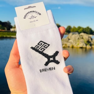 Puede incluir: Calcetines blancos con un diseño de llave negra y la palabra "BREMEN" impresa. La etiqueta muestra "SOUVENIRCLUB" y la talla 42-46. Los calcetines se muestran sobre un fondo de agua y cielo azul.