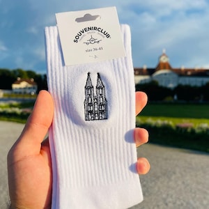 Könnte beinhalten: Weiße gerippte Socken mit einem schwarz gestickten architektonischen Design. Die Socken haben ein Etikett mit der Aufschrift "SOUVENIRCLUB" und "Größe 36-41". Die Socken werden vor einem Gebäude und blauem Himmel gehalten.