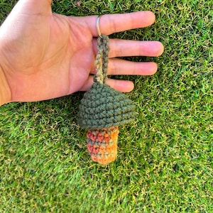 May include: A crocheted mushroom keychain with a green cap and orange and brown stem. The keychain is hanging from a silver ring.