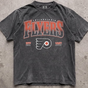 Op de afbeelding: Een antracietgrijs t-shirt met korte mouwen en de tekst Philadelphia Flyers in een rood en zwart verlooplettertype. Onder de tekst staat het Flyers-logo, het woord EST en het jaartal 1967 in het rood. Het shirt heeft een ronde hals en een vervaagde look.