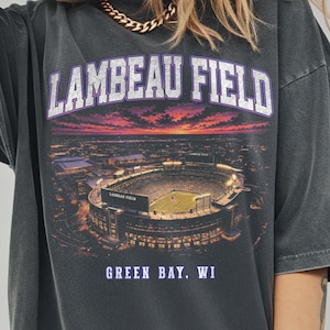 May include: A charcoal grey oversized t-shirt featuring a graphic print of Lambeau Field in Green Bay, WI. The design includes the text LAMBEAU FIELD in a purple-outlined font above a sunset stadium view and GREEN BAY, WI in purple text below.