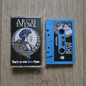 Op de afbeelding: Een blauwe cassette en hoesje met de band Arch Enemy. De hoes heeft een zwarte achtergrond met een afbeelding van een vrouw met blauw haar en vleugels. De cassette is blauw met zwart-witte afbeeldingen.