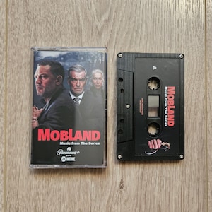 Könnte beinhalten: Eine schwarze Kassette und ihre Hülle werden gezeigt. Die Hülle zeigt den Titel "Mobland" in Rot, mit Bildern von Schauspielern. Die Kassette selbst ist schwarz, mit dem Titel "Mobland" in roter und weißer Schrift.