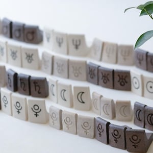 May include: A small bonsai tree in a round, brown ceramic pot sits on a cork base. A winding path of small, rectangular tiles with astrological symbols leads away from the plant. The tiles are in shades of brown, white, and gray.