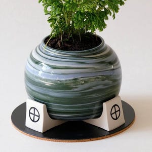 May include: A small, round ceramic planter with a green and blue marbled design, filled with a leafy green plant. The planter sits on a black coaster supported by a white stand with a geometric design.