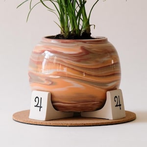 May include: A decorative ceramic planter with a swirling orange and brown pattern, holding a green plant with long, thin leaves. The planter sits on a white stand with the number "24" printed on it, placed on a round cork base.