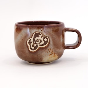 May include: A brown ceramic mug with a handle and a decorative emblem. The mug has a rounded shape and a dark brown exterior with lighter brown and beige accents. The emblem is a lighter brown color.