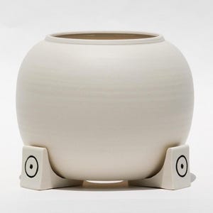 May include: A cream-colored ceramic vase with a rounded body and a wide opening. The vase rests on three square, off-white supports, each featuring a black circle design. The overall design is minimalist and modern.