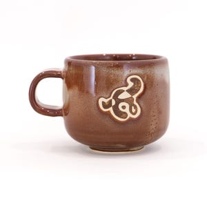 May include: A brown ceramic mug with a rounded shape and a handle. The mug features a cream-colored, abstract design on the front. The mug is set against a white background.