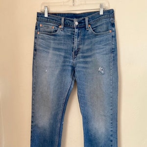 May include: Light blue denim jeans with a straight leg cut, featuring a faded wash and distressed details. The jeans are hanging on a white hanger, showcasing the classic five-pocket design and a button and zipper closure.