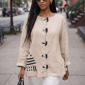 May include: Beige linen jacket with black toggle closures and long sleeves. The jacket features a black and white geometric design on the lower left side. The person is wearing white pants and holding a black handbag.