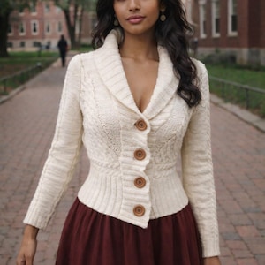 May include: Cream-colored cable knit cardigan with a shawl collar and wooden buttons. The long-sleeved sweater is paired with a burgundy skirt. The outfit is worn outdoors on a brick pathway.