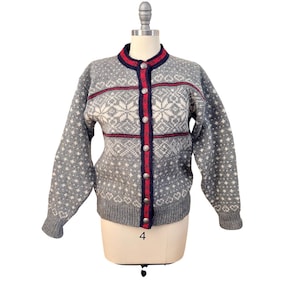 May include: A gray cardigan sweater with a snowflake and star pattern. The sweater has a round collar and is buttoned down the front with silver buttons. Red and navy blue stripes accent the collar and front. The sleeves have a heart pattern.