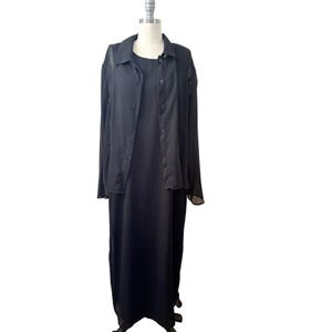 May include: A black dress and jacket set displayed on a mannequin. The dress is a long, solid black, sleeveless design. A sheer, button-down jacket with long sleeves is layered over the dress. The outfit is suitable for formal occasions.
