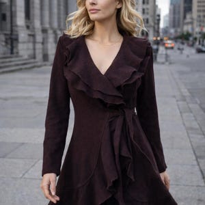 May include: A dark burgundy wrap coat with a ruffled collar and waist tie. The long-sleeved coat has a flared skirt and is worn over black pants. The woman is walking on a city sidewalk.