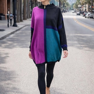 May include: A color-blocked tunic sweater in shades of purple, teal, and black. The sweater has a high neckline with gold buttons and is paired with black leggings and black flats. The outfit is worn outdoors on a city street.