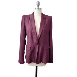 May include: A plum-colored blazer with a single-button closure and two front pockets. The blazer is displayed on a mannequin against a white background. The jacket has a classic lapel and long sleeves.