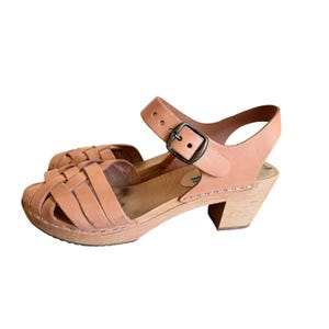 May include: A pair of light brown leather sandals with a woven design on the front. The sandals have a wooden sole and a small heel. The ankle strap features a metal buckle. The sandals are designed for casual wear.