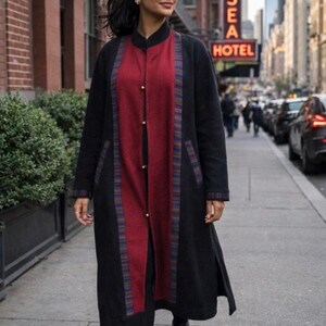 May include: A woman wearing a long black coat with a red panel down the front, accented with gold buttons and colorful trim. The coat is worn over black clothing and black boots. The background shows a city street with a hotel sign.