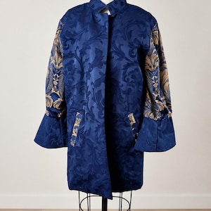 May include: A long-sleeved coat featuring a deep blue base with a tonal damask pattern. The sleeves are accented with a contrasting gold and blue ornate floral print. The coat includes a high collar, a front button closure, and side pockets.