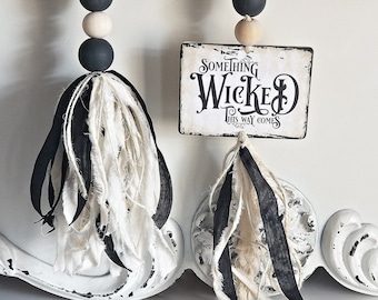 May include: Decorative wooden bead garland with black, white, and natural wood beads. Each strand features a tassel made of black and white fabric strips. One strand has a sign that reads "SOMETHING WICKED THIS WAY COMES."