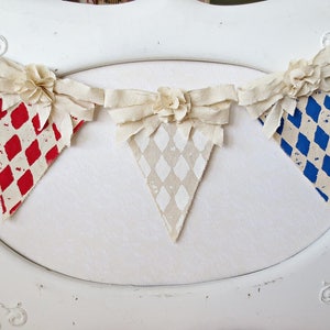 May include: A white fabric banner with three triangular flags. The flags are decorated with red and white checkered, white and gray checkered, and blue and white checkered patterns. The banner is decorated with white fabric flowers.