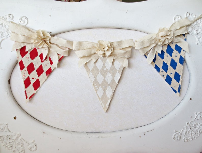 May include: A white fabric banner with three triangular flags. The flags are decorated with red and white checkered, white and gray checkered, and blue and white checkered patterns. The banner is decorated with white fabric flowers.
