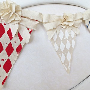 May include: A fabric banner with three triangular flags. Each flag is decorated with a different diamond pattern in red, white, and blue. The flags are connected by white fabric bows with large white fabric flowers.