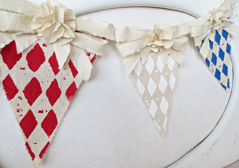 May include: A fabric banner with three triangular flags. Each flag is decorated with a different diamond pattern in red, white, and blue. The flags are connected by white fabric bows with large white fabric flowers.