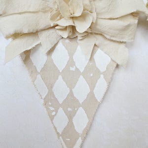 May include: A beige fabric triangle banner with a white diamond pattern. The banner is decorated with a large white fabric flower.